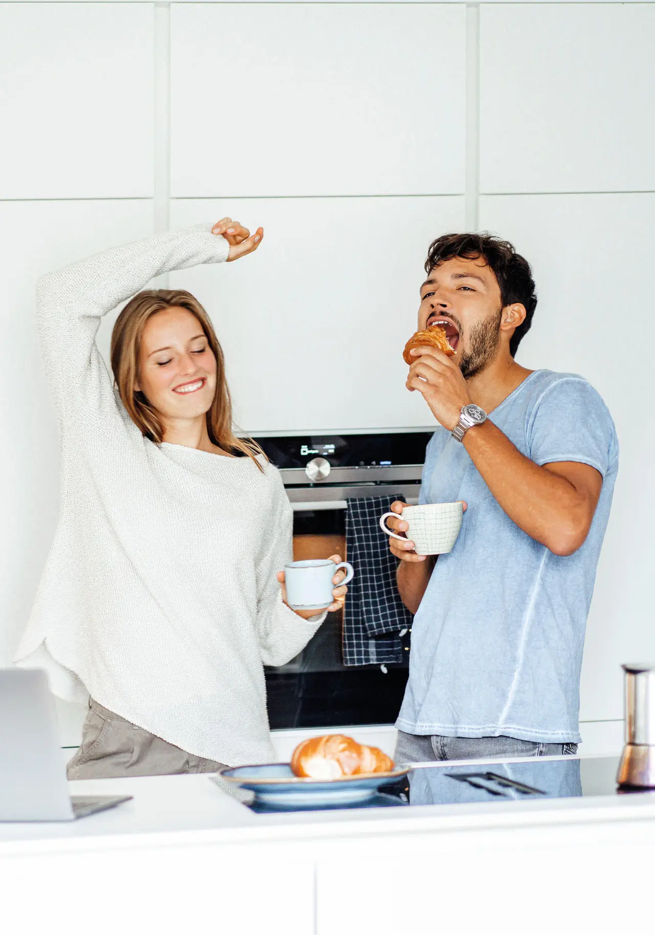 Junges Paar genießt Kaffee und Croissants in moderner Küche, fröhliche Stimmung am Morgen mit heller Kücheneinrichtung und Frühstücksszene.